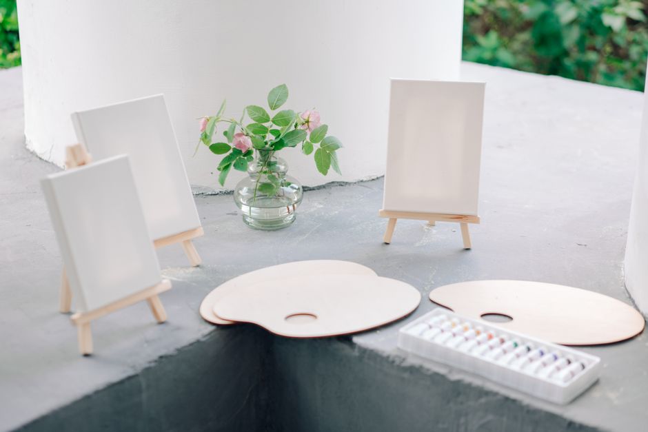 Art supplies setup with blank canvases on easels, paint palettes, and a jar of flowers.